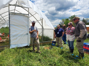 A farmer stands in front of a chicken schooner, speaking to a group of veterans. 